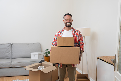 Happy man holds packed shipping boxes standing in newly renovated living room parcels with items for transportation by service and moving to new apartment