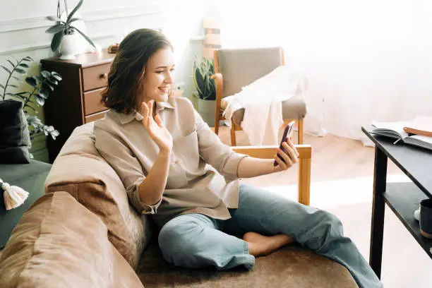Virtual Greetings. Smiling young woman engages in a video call, warmly waving her hand to say hello or hi, creating a welcoming atmosphere in the cozy living room setting