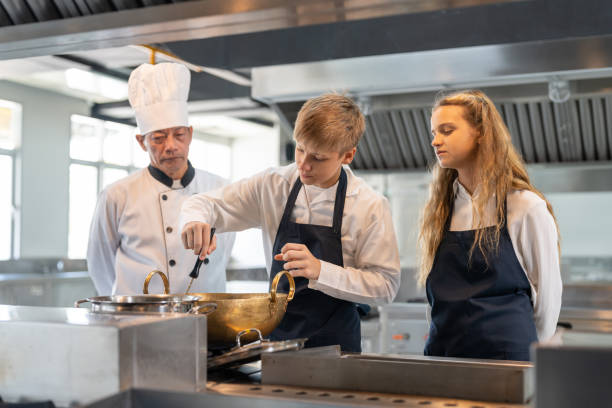 학교 부엌에서 수업에서 요리를 공부하는 학생 소년과 소녀의 그룹. 아이들에게 요리를 가르치는 수석 아시아 교사 요리사. 교육 개념 - chef trainee cooking teenager 뉴스 사진 이미지