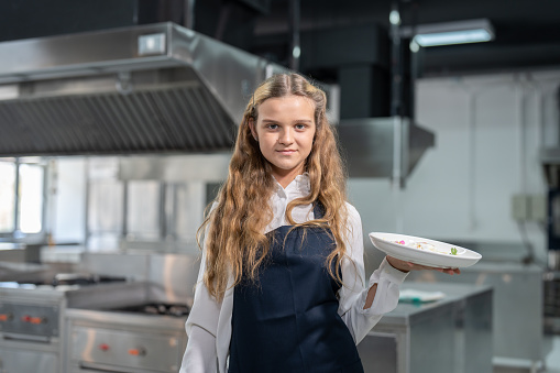 Portrait of student girl junior chef wearing apron holding her appetizer food in dish while studying cooking in kitchen room in class at school