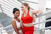 Two smiling females wearing fitnesswear standing on a bridge. Positive plus-size women relaxing during workout outdoors.