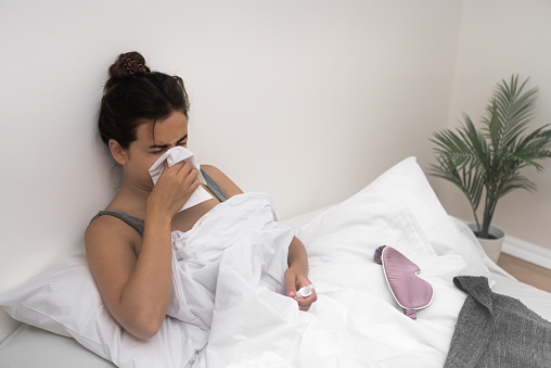 A unwell young lady, nose red and sniffles, uses tissues and clutches a bottle of medicine while sitting in bed, battling illness.