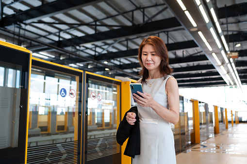Mature Asian business woman using smart phone to do online banking while waiting for mass rapid transit (MRT) on railway platform to travel for work.