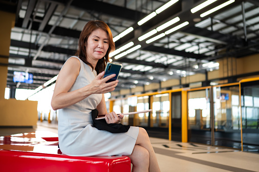 Mature Asian business woman using smart phone to do online banking while waiting for mass rapid transit (MRT) on railway platform to travel for work.