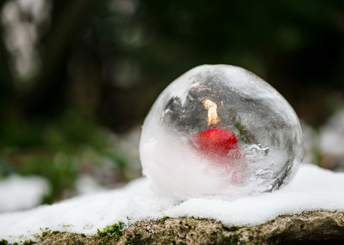Ice globe lantern with red candle burning inside in the evening garden. Winter craft and Ice art or outdoor decoration concept.
