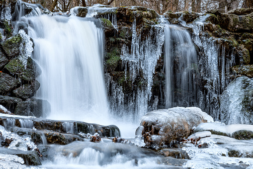 Icicles hang from the side of a waterfall, creating a stunning winter scene. The waterfall is located in a mountainous area, and the rocks are covered in ice. Hungary