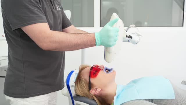Female patient in protective red glasses at UV teeth whitening procedure in the stomatology clinic. Dentist starting bleaching teeth procedure with young woman. Modern cosmetic dentistry.