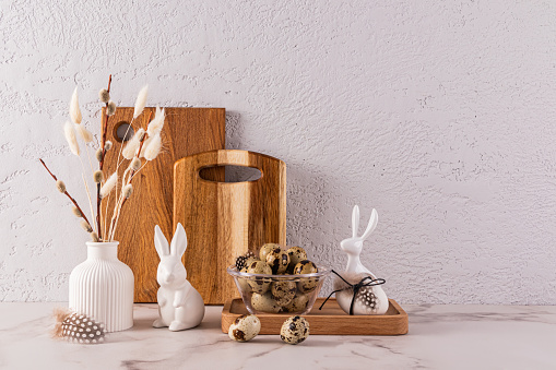 Stylish eco-friendly background for Easter holiday with ceramic Easter rabbit figurines, vase with willow twigs, bowl with quail eggs