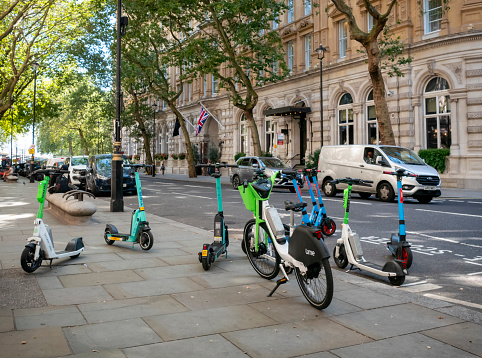 A collection of various rental electric scooters and an electric bike scattered about on a pavement in Northumberland Avenue in the City of Westminster, London. The group includes bikes owned by Dott, Lime and Tier. Only a few of the scooters are correctly parked in a designated bay.