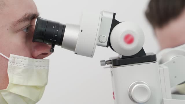 Close-up of male scientist in protective face mask using microscope in pharmaceutical laboratory. Microbiologist performs microscopic tests of vaccine. Scientist studies soil using microscope in lab.