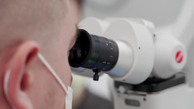 Close-up of male scientist in protective face mask using microscope in pharmaceutical laboratory. Microbiologist performs microscopic tests of the vaccine. Scientist studies research samples in lab.