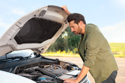 Upset man looking under hood of broken car outdoors