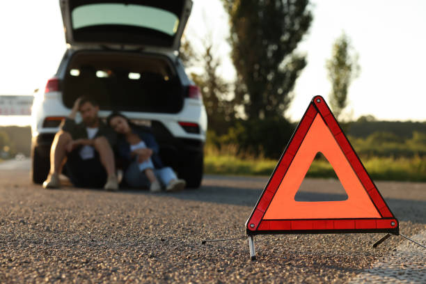 istock Warning triangle and couple sitting near broken car on roadside, selective focus 1932631775