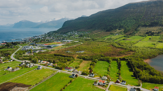 Drone high-angle photo of vehicle passing the highway toad through the authentic buildings, highway, fjord and mountains around during summertime