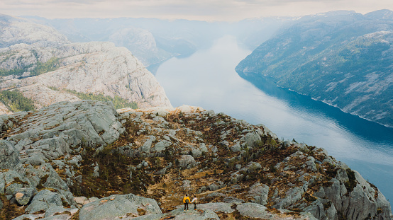 Done high-angle photo of heterosexual couple of backpackers getting to the edge of the cliff admiring beautiful landscape of mountain fjord during sunrise in South Norway, Scandinavia