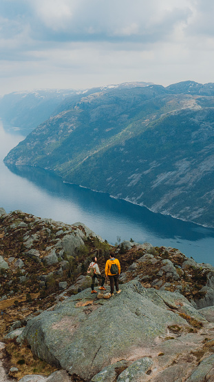 Done high-angle photo of heterosexual couple of backpackers getting to the edge of the cliff admiring beautiful landscape of mountain fjord during sunrise in South Norway, Scandinavia