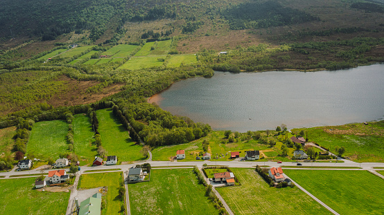 Drone high-angle photo of vehicle passing the highway toad through the authentic buildings, highway, green forest, lake, sea and mountains around during summertime