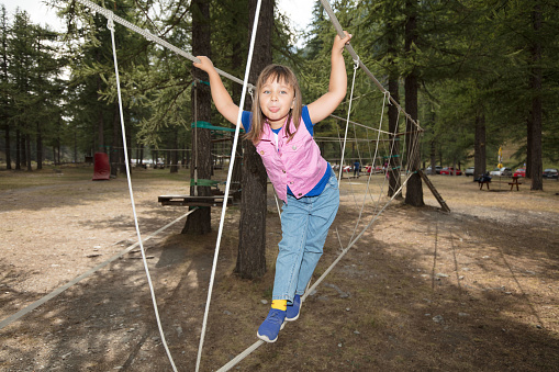 A little girl plays in the adventure park, amidst big larches. She walks on a rope, balancing with her hands.