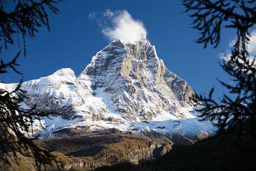 An early autumn view of the Italian side of the Matterhorn or Monte Cervino, resembling a granite pyramid. A plume of clouds envelops the summit.