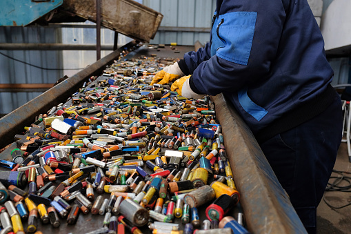 A worker in blue attire and protective gloves sorts through a heap of assorted spent batteries on a conveyor belt at a recycling plant.