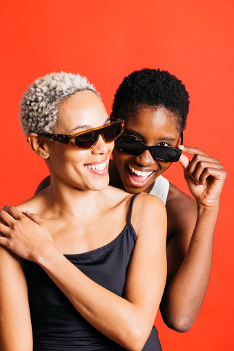Portrait of a cheerful young female couple, wearing sunglasses, together in front of a red background at studio. Two women together at studio.