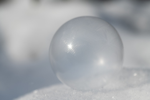 Close-up of a delicate soap bubble that is frozen. The background is light-coloured. The soap bubble lies on snow. The sun is shining.