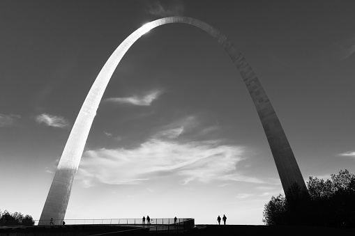 St. Louis, Missouri - USA: The Gateway Arch at sunset in black and white. Sun reflecting on left half of arch with dark sky and clouds. Silhouettes of couple walking on horizon against sky.