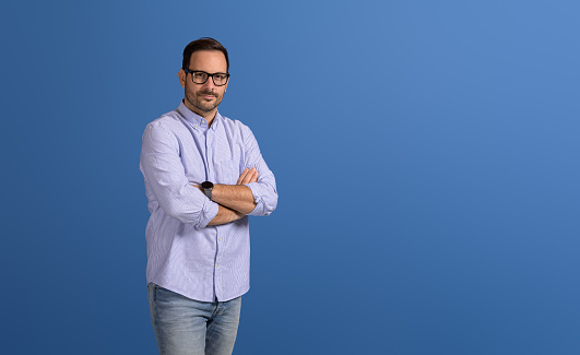 Portrait of confident serene young businessman with arms crossed posing on isolated blue background