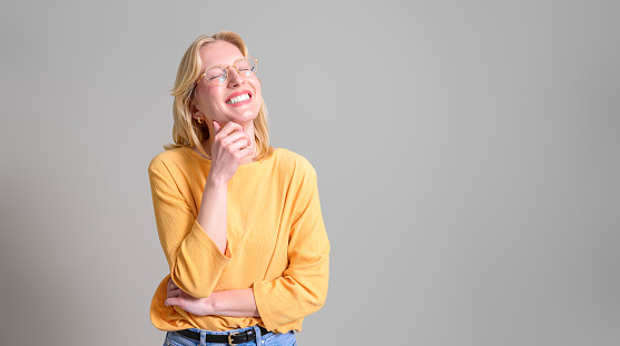 Smiling blond businesswoman with eyes closed and hand on chin standing isolated on white background