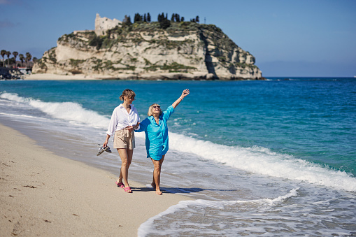 Senior woman and her mature daughter enjoying spring day in Calabrian town of Tropea. They are walking on the beach, holding hands, talking and having fun. Springtime, off-season vacations day in Catania, Italy.\nShot with Canon R5