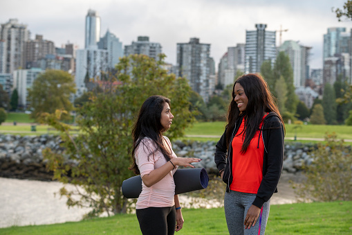 Young women walk home from yoga class together through city park, skyline and harbour in distance