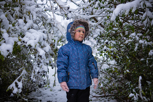 Little beautiful girl in winter clothes standing alone in the middle of a snowy forest. Girl in jacket with fur hood posing in winter forest. Shrubs and trees are covered with snow
