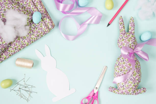 istock Easter flat lay. Soft decorative toy Easter Bunny made of cotton fabric in small flower, decorated with pink satin ribbon bow. Supplies needed to make craft. Layout on blue background. Top view. DIY 1933286182