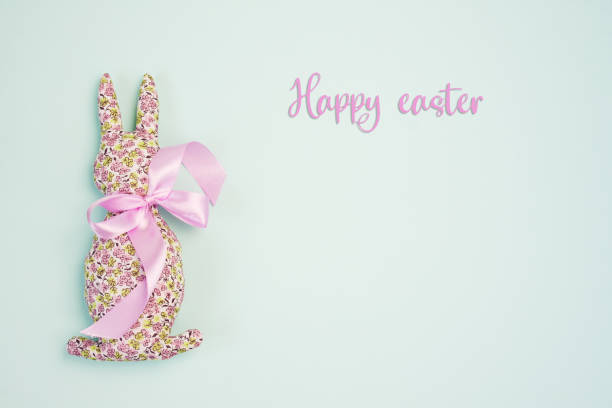 Happy easter. Decorative handmade Easter Bunny soft toy made of cotton fabric, decorated with pink satin ribbon. Isolated on blue background. Mockup for greeting card. Space for text. top view