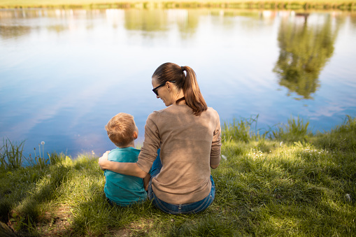 Lovely Asian mother and adorable baby boy feeding fish in urban garden together. Little toddler boy feeding fish in pond with his mother. Young baby animal experience outdoor learning family relation concept.