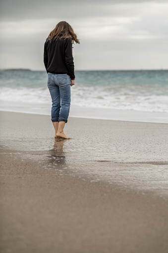 Full-length rear view of long-haired woman standing barefoot on the water's edge. Vertical composition, copy space.