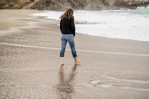 Adult woman walking relaxed on a beach in winter observing the effects of the water flowing on the foreshore. Full-length rear view, horizontal composition, copy space.