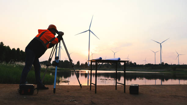 ingegnere con giubbotto di sicurezza arancione che utilizza il lavoro della telecamera di rilevamento nel campo della fattoria dei mulini a vento - engineer environment orange manual worker foto e immagini stock