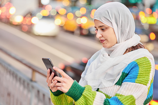 Portrait of a young Muslim woman wearing hijab using a smartphone. She is on the street in the city. Dressed in casual clothes with hijab on her head.