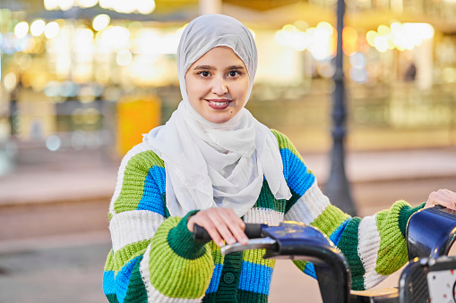 Young Muslim woman renting a bicycle in the city. She is smiling and looking at the camera. She is wearing hijab on her head and casual clothes.