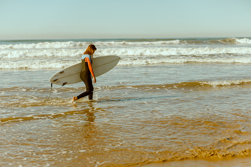 Happy female surfer in wetsuit with his surfboard entering out of sea after surfing on waves