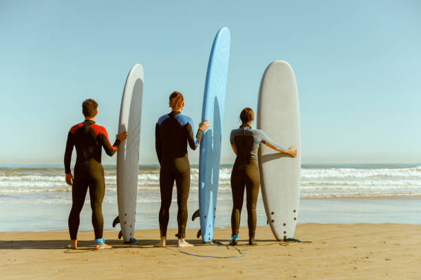 istock Back view of surfers in wetsuits standing with surfboards and looking on ocean 1933365862