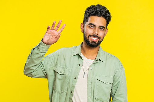 Young Indian man smiling friendly at camera, waving hands gesturing hello greeting or goodbye welcoming with invitation hospitable expression. Arabian Hindu guy isolated on yellow studio background