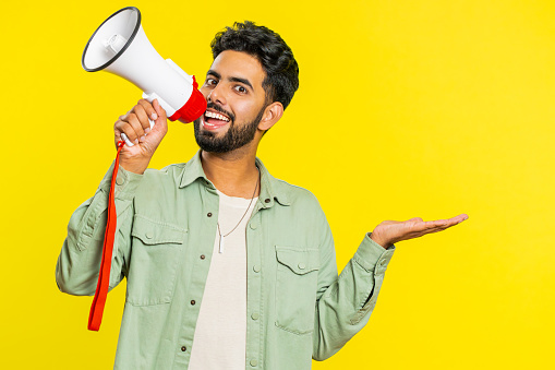 Indian young man talking with megaphone, proclaiming news, loudly announcing advertisement discounts sale, using loudspeaker to shout speech. Happy Arabian guy isolated on studio yellow background