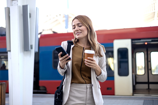 A young businesswoman is using her mobile phone and carrying a cup of coffee as she uses the train to travel to work in a morning