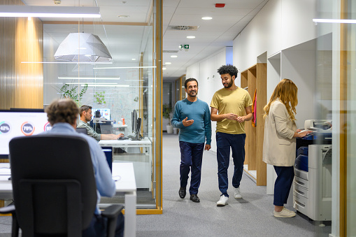 Diverse coworkers chatting and walking in crowded office corridor, medium shot view behind glass.