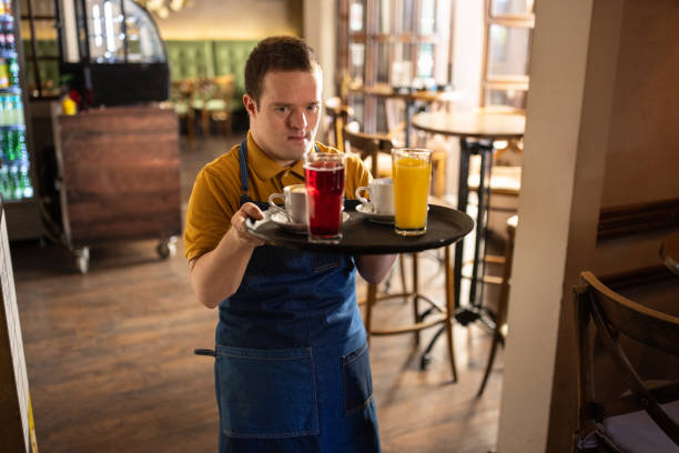 Charming man with down syndrome working as a waiter in cafe, he is carrying drinks on a tray.