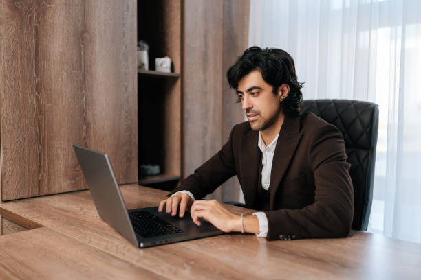 istock Handsome bearded businessman sitting at workplace by window, working on laptop, looking at screen, typing messages. Businessman dressed in suit 1933646399