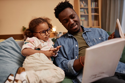 Medium long shot of little African American girl wearing glasses at home with dad reading stories