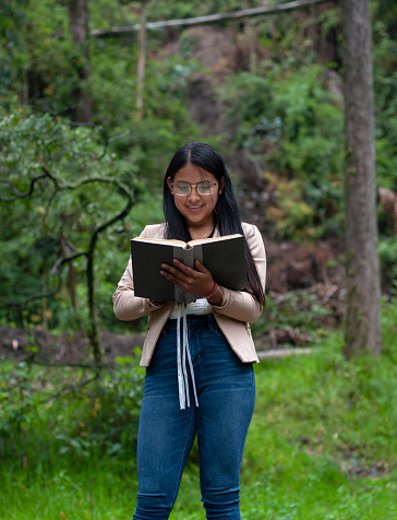 latin girl walking through a forest surrounded by vegetation and trees while excitedly reading a book.